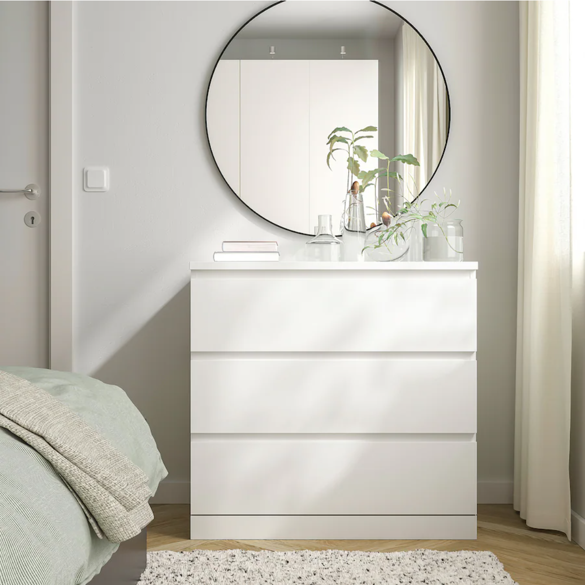 A white MALM chest of drawers with a round mirror above it, placed against a wall with a bed and window in the background.