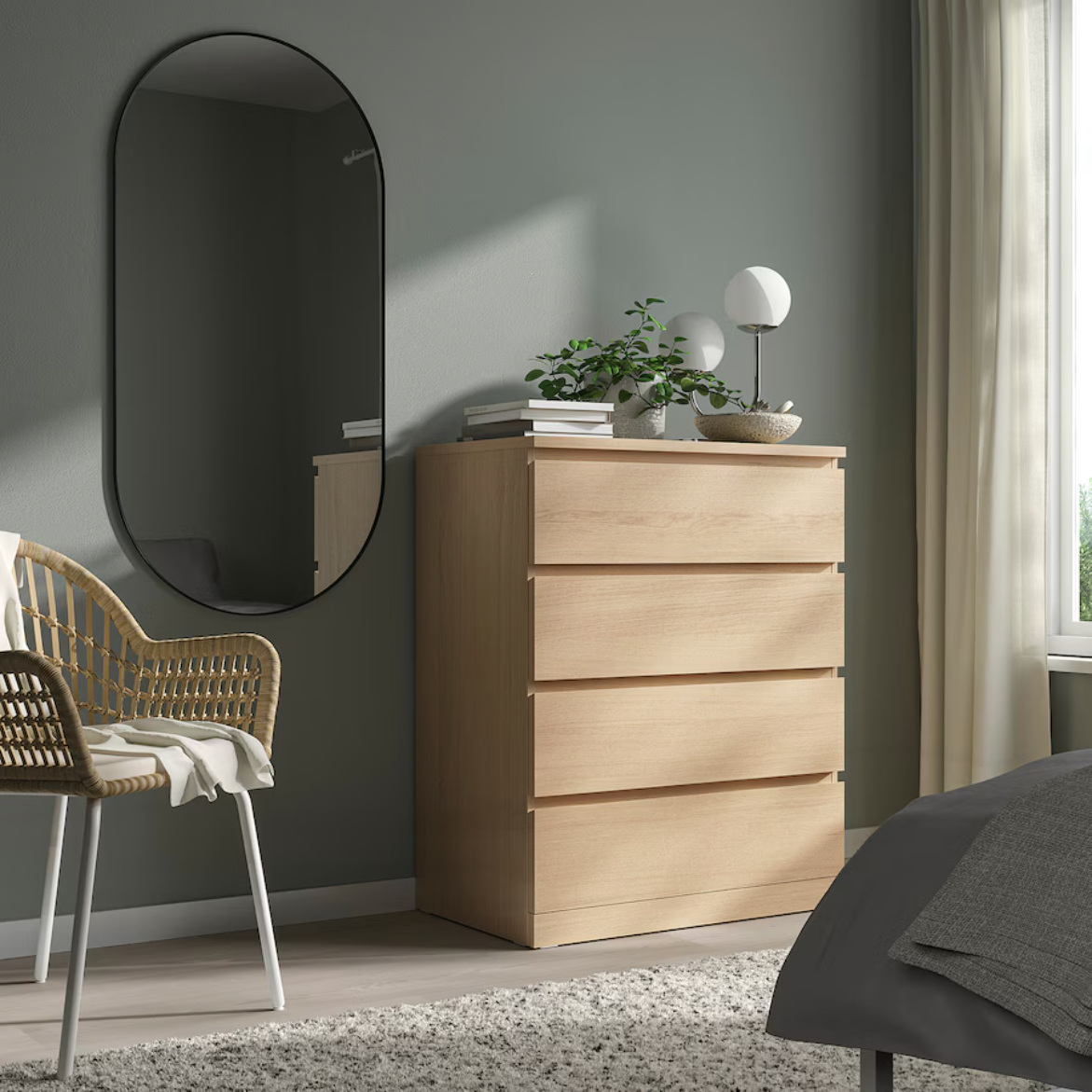 A white stained oak veneer chest of four drawers placed against a wall with a round black mirror above a chair to the left. The room is softly lit with natural light and has a serene atmosphere.